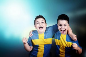 Two adorable boys soccer fans with flag of Sweden on t-shirt, embracing celebrate the victory of his team. Happy and shout. Stadium lights in the background. Blue background. Landscape orientation.